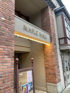 A photo of the exterior of Maple Hall in La Conner. The exterior is mainly brick with marquee-style lightbulbs illuminating the words "Maple Hall".