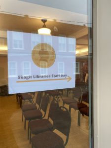 Photo of a window with a sign on it saying "Skagit Libraries Staff Day" and an arrow pointing right. You can see chairs arranged in a row through the glass.