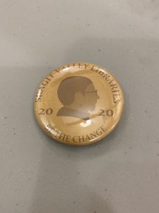 Photo of a golden button pin with drawing of Nancy Pearl on it and the words Skagit Valley Libraries 2020 Be The Change.