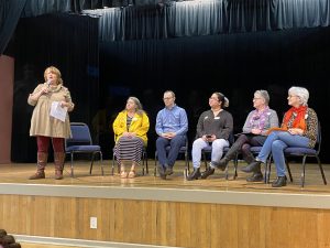 A female facilitator speaks to a staff member audience as the Skagit County Directors and Acting Directors sit on the stage on chairs and look on.