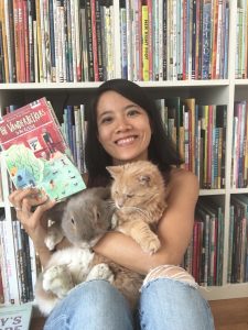 Photo of Karina Yan Glaser sitting in front of a bookshelf holding a book, a large grey bunny, and an orange cat. She is smiling.